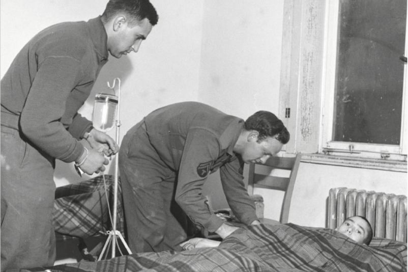 Das Bild zeigt eine Szene in einem Krankenzimmer oder einem provisorischen medizinischen Bereich. Zwei Männer in Uniform kümmern sich um eine liegende Person, die unter einer karierten Decke liegt. Der Mann auf der rechten Seite beugt sich über den Patienten und scheint seine Vitalzeichen zu überprüfen oder ihm zu helfen, während der andere Mann auf der linken Seite eine medizinische Infusion vorbereitet. Im Hintergrund ist ein Fenster mit einem alten Rahmen zu sehen, und unter dem Fenster steht ein Heizkörper. Die Umgebung wirkt schlicht und funktional, möglicherweise in einem militärischen oder Notfalleinsatz-Kontext. Die Körperhaltung der beiden Männer deutet darauf hin, dass sie sich fürsorglich und professionell um die liegende Person kümmern.